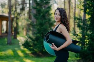 zijwaarts schot van mooie slanke vrouw met perfect figuur, golvend donker haar, houdt opgerolde karemat en fitnessbal vast, poseert buiten tegen een wazige bosachtergrond, heeft aerobicsoefeningen in de ochtend foto