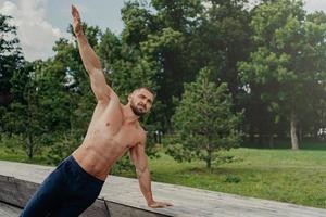 sterke jonge man staat in zijplank op één arm, vindt balans, poseert in het park in de buurt van bomen, beoefent yoga buiten, leidt een actieve gezonde levensstijl, heeft een gespierd sterk lichaam. gemotiveerde bodybuilder foto