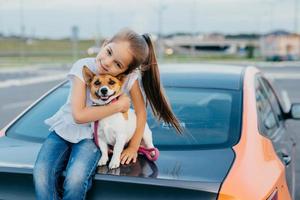 foto van vrolijk klein vrouwelijk kind heeft een paardenstaart omhelst met liefde haar favoriete huisdier, poseert in de kofferbak van de auto, speelt samen, loopt in de open lucht, geniet van het samenzijn. kinderen en dieren concept