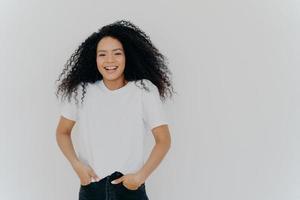 halflange foto van knappe glimlachende vrouw lacht om grappige grap, heeft plezier, houdt beide handen in de spijkerbroekzakken, draagt een wit t-shirt, heeft pluizig krullend haar, poseert binnen, lege ruimte aan de rechterkant