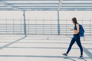 volledig portret van sportieve actieve vrouwelijke tiener of jongere, draagt top, legging en sneakers, draagt rugzak, geniet van aangename muziek in afspeellijst, gebruikt moderne mobiele telefoon en oortelefoons foto