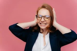 gefrustreerde jonge zakenvrouw heeft hoofdpijn, negeert hard geluid, bedekt oren met handen, klemt tanden op elkaar, heeft ogen gesloten, draagt elegante zwart-witte formele kleding, geïsoleerd op roze achtergrond foto