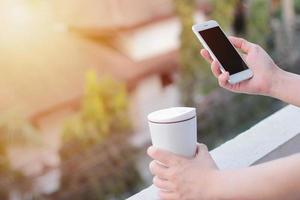 close-up van vrouwenhanden die smartphone gebruiken en thermosmok vasthouden, kopieer ruimte met zonlicht foto