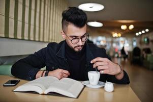 arabische man draagt een zwarte spijkerjas en een bril die in café zit, boek leest en koffie drinkt. stijlvolle en modieuze Arabische modelman. foto