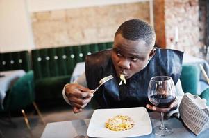 afrikaanse man in zwarte traditionele kleding zit in restaurant en eet pasta en drinkt wijn. foto