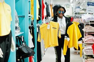 Afro-Amerikaanse vrouwen in trainingspakken en zonnebrillen winkelen bij sportkleding winkelcentrum tegen planken. ze koos geel t-shirt. sport winkel thema. foto