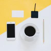 creatieve platliggende foto van werkruimtebureau met smartphone, koffie en tag op gele en witte achtergrond, minimaal gestyled