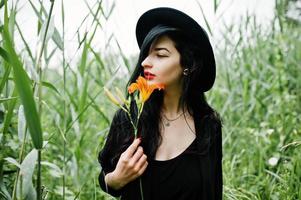 sensueel meisje helemaal in het zwart, rode lippen en hoed. goth dramatische vrouw houdt oranje leliebloem op gemeenschappelijk riet. foto