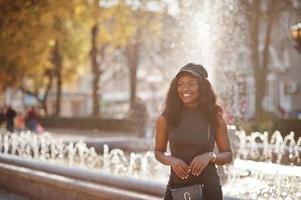 stijlvol Afrikaans Amerikaans meisje in grijze tuniek, crossbody tas en pet gesteld op zonnige herfstdag tegen fonteinen. afrika model vrouw. foto