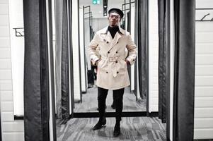 stijlvolle casual Afro-Amerikaanse man bij zwarte baret en witte trenchcoat bij paskamerkledingwinkel. foto