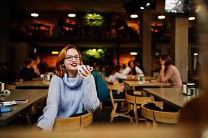 vrolijke jonge mooie roodharige vrouw in glazen zittend op haar werkplek op café en koffie drinken. foto