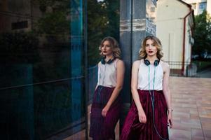 modieuze en mooie blonde model meisje in stijlvolle rood fluwelen velours rok, witte blouse, geposeerd met telefoon en koptelefoon tegen ramen van stadsgebouw. foto