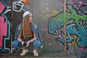 stijlvolle casual hipster meisje in pet en spijkerbroek dragen luisteren muziek van koptelefoon van mobiele telefoon tegen grote graffiti muur met bom. foto