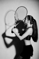 zwart-wit portret van een mooie jonge vrouw in sportkleding die een tennisracket vasthoudt terwijl hij tegen een witte achtergrond staat. foto