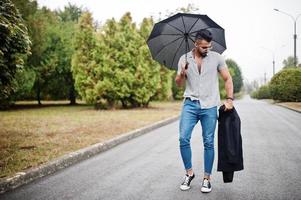 modieuze lange Arabische baard man slijtage op shirt, jeans en zonnebril wandelen in het park met paraplu en jas bij de hand. foto