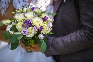 de bruid legde haar handen op de schouders van de bruidegom. bruid met een boeket van violette en witte rozen knuffels en kus de bruidegom in de natuur. foto