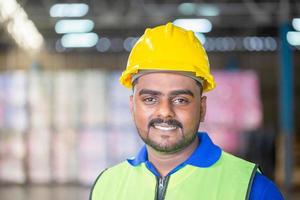 portret van werknemer met veiligheidshelm en veiligheidsvest in het magazijn, vrolijke man die naar camera kijkt met onscherpe achtergrond en kopieerruimte foto