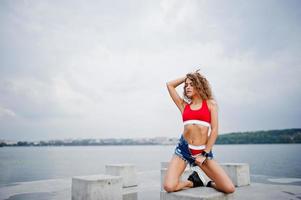 sexy krullend model meisje in rode top, jeans denim shorts en sneakers geposeerd op stenen kubussen tegen bewolkte hemel en meer. foto