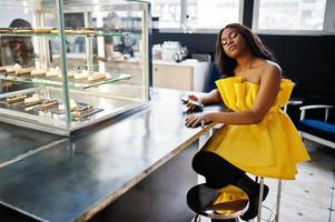 stijlvolle Afro-Amerikaanse vrouw in gele jurk poseerde in café met mobiele telefoon bij de hand. foto