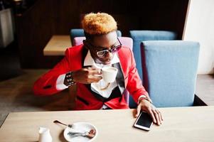 mode Afro-Amerikaanse man model in rood pak, met hoogtepunten haar en zonnebril zittend in café met koffie. foto
