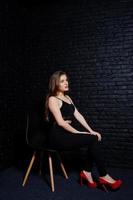 knappe brunette meisje slijtage op zwarte en rode hoge hakken, zittend en poseren op stoel in studio tegen donkere bakstenen muur. studiomodel portret. foto