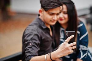 indiase paar poseerde buiten, zittend op een bankje samen en kijken naar mobiele telefoon, selfie maken. foto