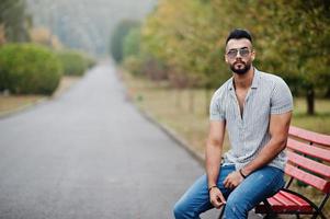 modieuze lange Arabische baard man slijtage op shirt, jeans en zonnebril gesteld op park op bankje. foto