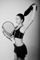 zwart-wit portret van een mooie jonge vrouw in sportkleding die een tennisracket vasthoudt terwijl hij tegen een witte achtergrond staat. foto