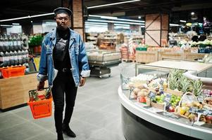 stijlvolle casual Afro-Amerikaanse man bij jeans jasje en zwarte baret met mand met ananas in fruit biologische sectie van supermarkt. foto