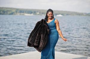 Afro-Amerikaanse donkere huid plus size model poseerde in een blauwe glanzende jurk en zwarte bontjas tegen de zeezijde. foto