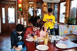 groep aziatische vrienden die in café zitten. gelukkige indiase mensen die samen plezier hebben, op de bank zitten en naar mobiele telefoons kijken. foto