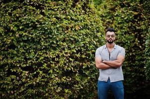 modieuze lange Arabische baard man slijtage op shirt, jeans en zonnebril gesteld op park tegen groen. foto