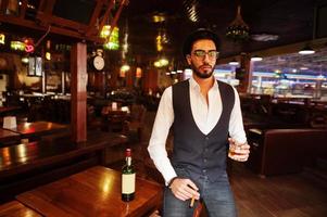 knappe goed geklede arabische man met glas whisky en sigaar poseerde in de pub. foto