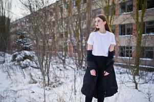 modieuze lange benen brunette model in lange zwarte mantel geposeerd buiten op winterdag tegen verlaten fabriek. foto