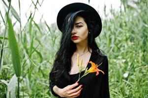 sensueel meisje helemaal in het zwart, rode lippen en hoed. goth dramatische vrouw houdt oranje leliebloem op gemeenschappelijk riet. foto