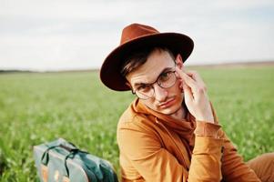 stijlvolle man met bril, bruine jas en hoed met tas op groen veld. foto