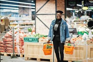 stijlvolle casual Afro-Amerikaanse man bij jeans jasje en zwarte baret met twee manden, wandelen en winkelen bij supermarkt. foto