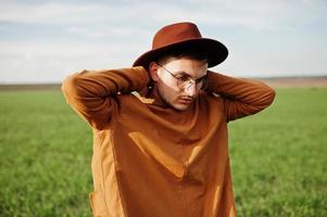 stijlvolle man in bril, bruine jas en hoed gesteld op groen veld. foto