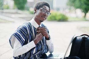 Afrikaanse man student in traditionele kleding en bril met rugzak, werken op laptop buiten. foto