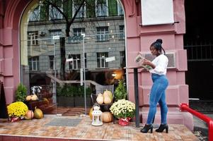 stijlvolle Afro-Amerikaanse vrouwen in witte blouse en spijkerbroek poseerden in de buurt van café met krant. foto