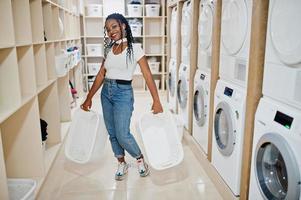 vrolijke afro-amerikaanse vrouw in de buurt van wasmachine die muziek luistert via oortelefoons van mobiele telefoon in de zelfbedieningswasserette. foto