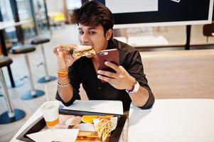 stijlvolle en grappige indiase man zit in fastfoodcafé en eet hamburger en maakt selfie via de telefoon. foto