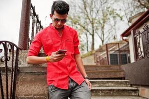 indiase man met rood shirt en zonnebril poseerde buiten en keek naar zijn telefoon. foto