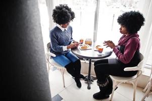 twee Afro-Amerikaanse vrouw met krullend haar draagt truien die binnen in café zitten met telefoons bij de hand. foto