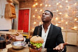 bid voor het eten. respectabele jonge Afro-Amerikaanse man in zwart pak zittend in restaurant met smakelijke dubbele hamburger en frisdrank. foto