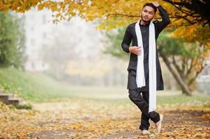 Indiase stijlvolle man in zwarte traditionele kleding met witte sjaal buiten poseerde tegen gele herfstbladeren boom. foto