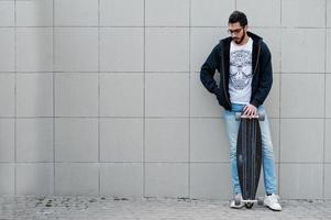 straatstijl Arabische man in brillen met longboard gesteld tegen grijze muur. foto