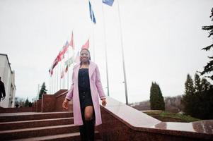 jonge stijlvolle mooie afro-amerikaanse vrouw in straat, gekleed in mode-outfit jas en bril, tegen vlaggen van verschillende landen van de wereld. foto