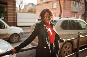 portret van een Afrikaanse vrouw met krullend haar, gekleed in modieuze zwarte jas en rode coltrui die buiten poseert. foto