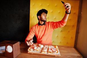 zelfverzekerde jonge indiase man in oranje trui zit bij pizzeria, eet pizza en maakt foto op zijn telefoon.